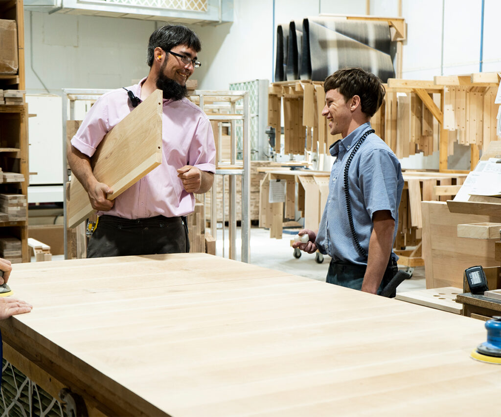 Two craftsmen discuss customizing a hardwood table inside an Amish workshop.