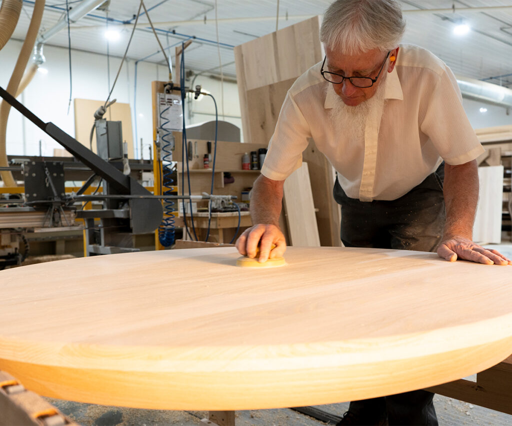 Woodworker hand-sanding a round hardwood tabletop in a small workshop.